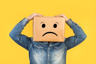 Kafası bir kutuda ve üzerine hüzün işareti çizilmiş bir adam ve sarı bir arka plan.
