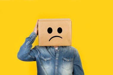 Kafası kutuda olan ve üzerine hüzün işareti çizilmiş bir adam. Sarı arkaplan