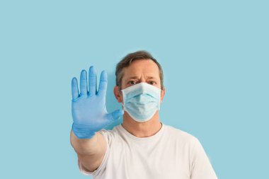 Açık mavi arka planda duran, tıbbi eldivenleri ve maskesi olan ve jest yapan bir adam.