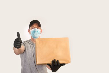 Eldivenleri ve maskesi olan bir teslimatçı. Elinde bir kutu var ve baş parmağı önde.