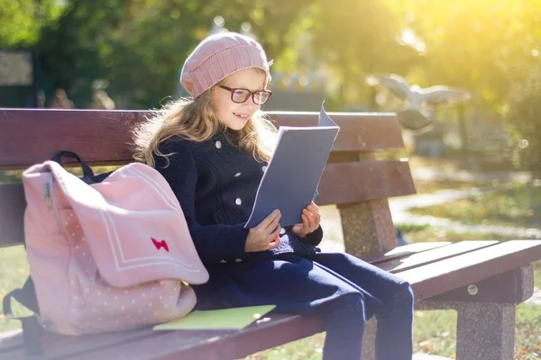 Mały uczeń szkoły podstawowej dziewczyna siedzi na ławce z plecakiem, patrzy na szkolnych książek i zeszytów. — Zdjęcie stockowe