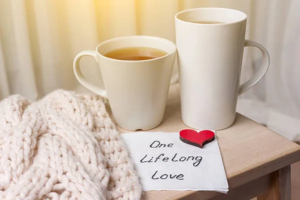 One long life of love is an abstract symbolic image. Couple of cups, background warm scarf, in home interior, napkin with text. — Stock Photo, Image