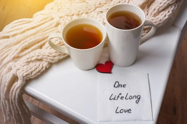One long life of love is an abstract symbolic image. Couple of cups, background warm scarf, in home interior, napkin with text. — Stock Photo, Image