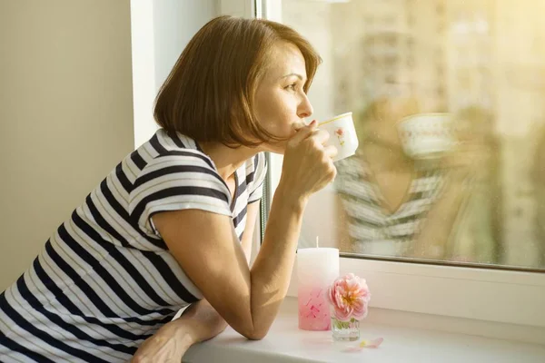 Mujer adulta bebe café, mira por la ventana en casa . — Foto de Stock
