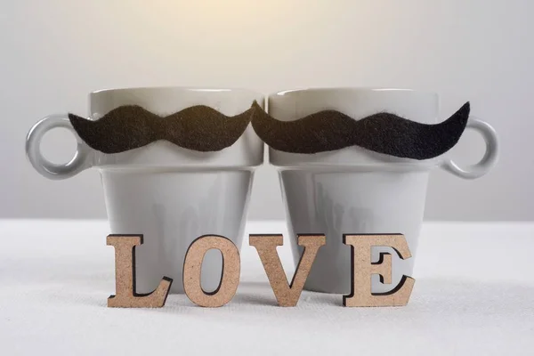Fondo romántico del día de San Valentín. Un par de tazas de bigotes negros - bigotes negros y una palabra de letras de madera - amor . — Foto de Stock