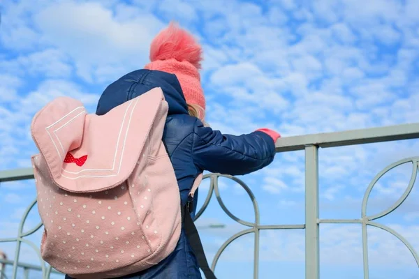 Vista posterior de niña con una mochila, en un sombrero de punto y chaqueta, fondo cielo azul con nubes . — Foto de Stock