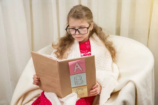 Mädchen mit Brille lernen, ein Buch mit dem Alphabet zu lesen. Hintergrund Stuhl im Wohnzimmer, Herbst-Winter-Saison. — Stockfoto