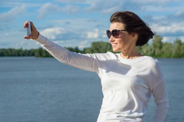 Bir kadın selfie smartphone kullanarak yapar. Arka plan mavi gökyüzü bulutlu, nehir.