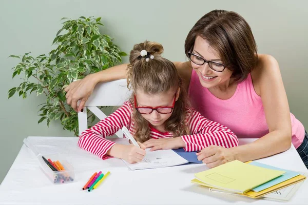 Madre amorosa ayudando a su hija alumna de la escuela primaria a escribir en su cuaderno, en casa en la mesa . — Foto de Stock