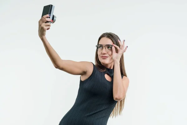 Zakenvrouw in een zwarte jurk en bril maakt selfie op een retro camera. — Stockfoto