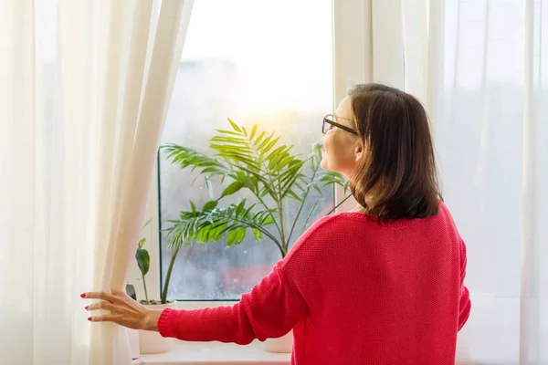 Vrouw kijkt uit het raam, de gordijnen opent. Uitzicht vanaf de achterkant — Stockfoto