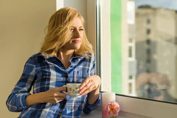Mujer rubia adulta bebe café, mira por la ventana en casa — Foto de Stock