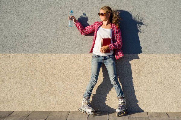 Girl teenager for an active lifestyle. Poses on the background of the wall is shod in the quad roller skates, holds a book and a bottle of water