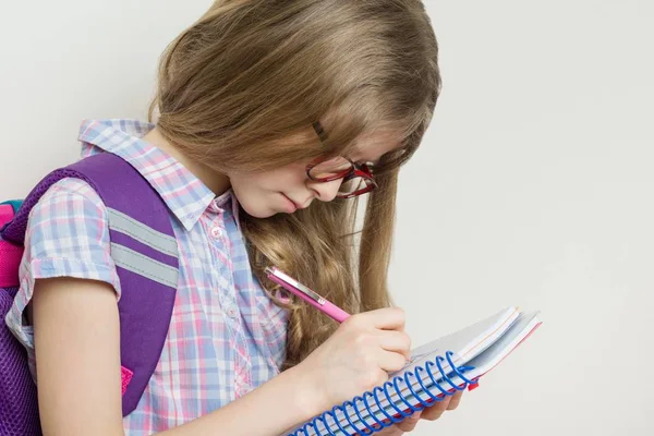 Niña estudiante de escuela primaria con gafas con una mochila escribiendo en su cuaderno . — Foto de Stock