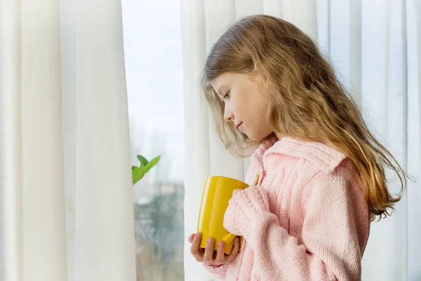 Девочка 7 лет блондинка с длинными волнистыми волосами в теплом трикотажном свитере держит чашку чая и смотрит в окно — стоковое фото