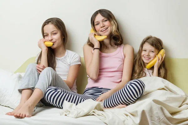 Hermanas niños en pijama juegan por la mañana en la cama. Mantenga los plátanos como teléfonos hablando y riendo —  Fotos de Stock