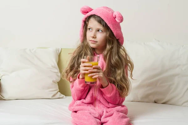 Μια κουκούλα pajama κορίτσι παιδί κάθεται στο κρεβάτι στο σπίτι και πίνει χυμό πορτοκάλι — Φωτογραφία Αρχείου