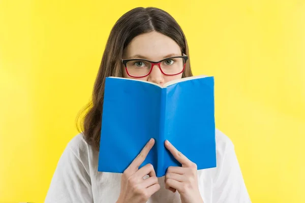 Closeup portret tienermeisje met bril kijkt uit over een boek, gele achtergrond — Stockfoto