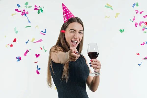 Mujer sonriente positiva en la fiesta de celebración con una copa de vino y en un sombrero de festival, Apunta con el dedo hacia ti —  Fotos de Stock