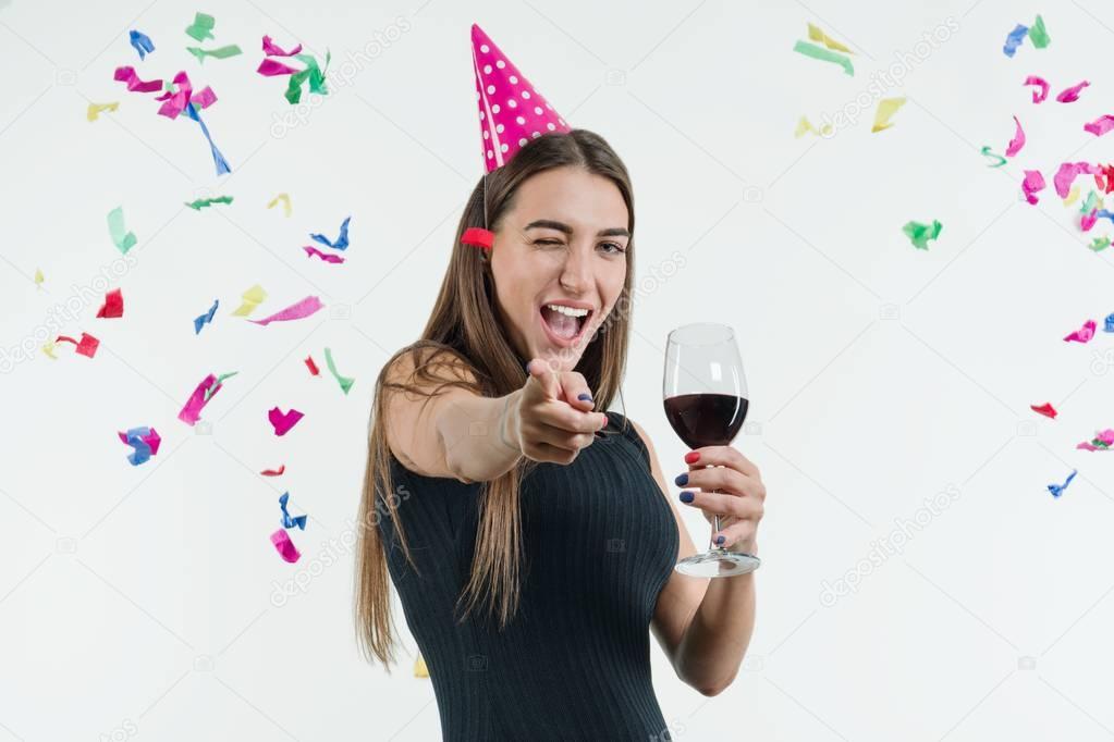 Positive smiling woman at celebration party with a glass of wine and in a festival hat, Point your finger at you