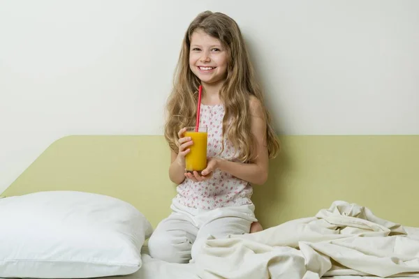 Сонная девушка 7 лет в пижаме, сидит в постели дома и пьет апельсиновый сок — стоковое фото