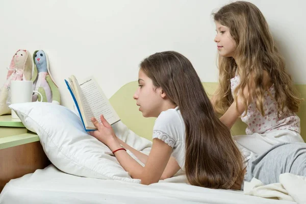 Дети читают книгу, лежащую дома в постели. Старшая девочка читает вслух младшей сестре — стоковое фото