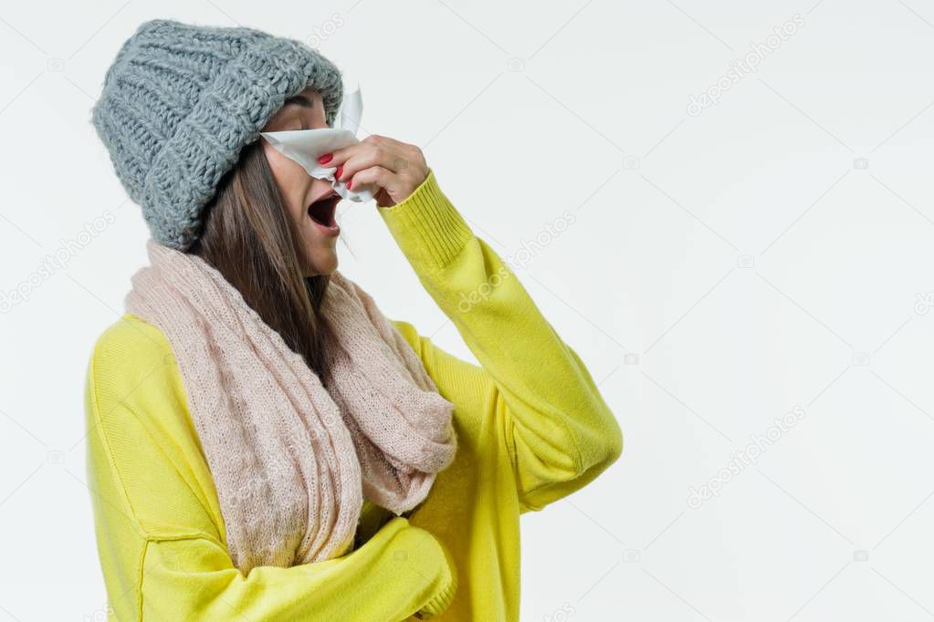 Woman in a sweater, knitted hat, scarf sneezes with a handkerchief. Season of the common cold, viruses, rhinitis.