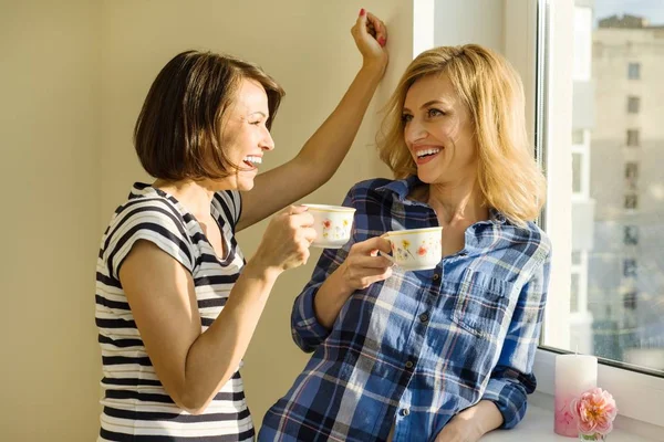 Adult women drink coffee, talk, laugh. Background home, solar window