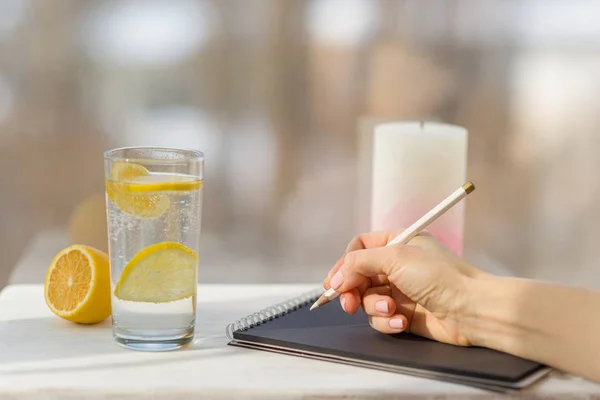 Mujer mano dibuja en cuaderno negro diseñador. Ventana de fondo, vaso de agua mineral con limón — Foto de Stock