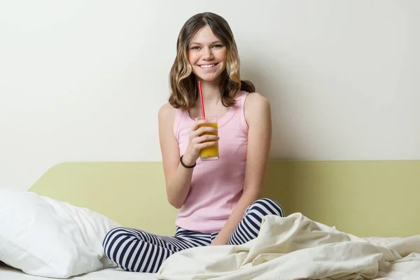 Улыбающаяся девочка-подросток 13 лет в пижаме, сидящая в постели дома и пьющая апельсиновый сок — стоковое фото