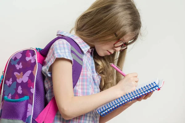 เด็กนักเรียนสาวในแว่นตาและกระเป๋าเป้สะพายหลังเขียนในสมุดบันทึกโรงเรียน . — ภาพถ่ายสต็อก