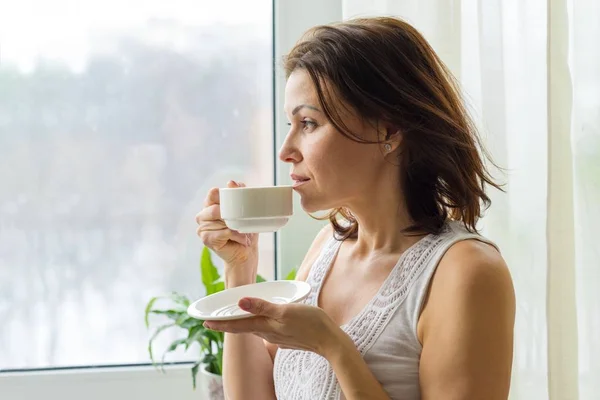 Mujer madura bebe café de la mañana y mira por la ventana en casa. Foto en perfil femenino grave — Foto de Stock