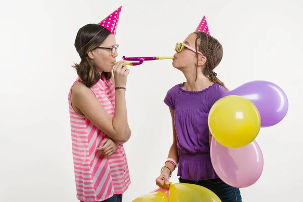 Tiener meisjes op een feestje. Meisjes op een witte achtergrond, in feestelijke hoeden, waait in de pijpen. — Stockfoto