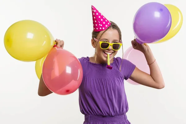 Veselá dospívající dívka 12,13 let, s balónky, v slavnostní klobouk, foukání potrubí na bílém pozadí, — Stock fotografie