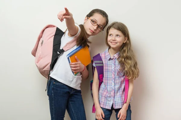 Duas crianças em idade escolar com mochilas fundo parede brilhante na escola, olhar e mostrar para o lado — Fotografia de Stock