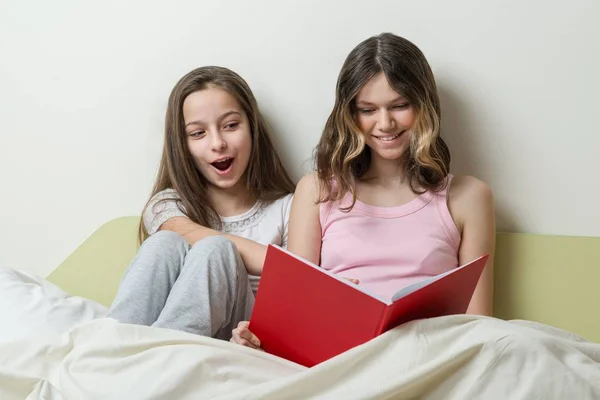 Dos adolescentes en pijama se sientan en la cama en casa y miran el libro . —  Fotos de Stock