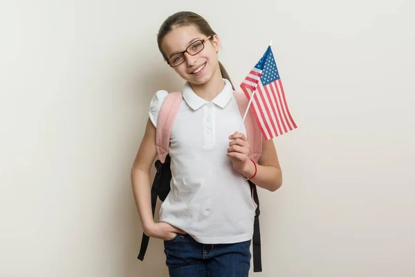 School child with a backpack holds the US flag, background bright wall in the school — Stock Photo, Image