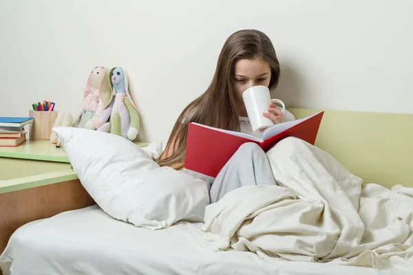 Dospívající dívka 10 let v pyžamu čte knihu a drží šálek čaje. — Stock fotografie