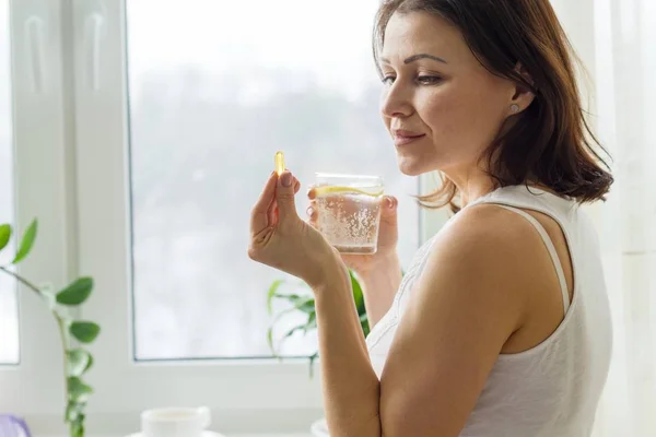 Kvinna tar p-piller med omega-3 och hålla ett glas rent vatten med citron. Bild på huset, på morgonen nära fönstret. — Stockfoto