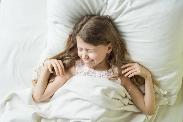 Goedemorgen, kind meisje blond met lang golvend haar wakker, ogen wrijft. Liggend op een kussen in bed, bovenaanzicht. — Stockfoto