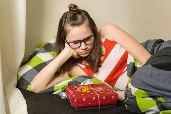 Triste chica adolescente decepcionado mirando regalo sentado en la cama en casa — Foto de Stock