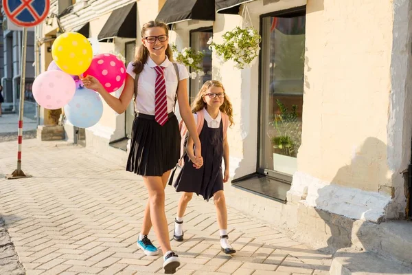 Duas meninas estão andando ao longo da rua com balões coloridos. Crianças de uniforme escolar, óculos, mochila — Fotografia de Stock