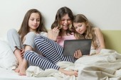 Három lány pizsamás ül az ágyban otthon érdeklődéssel nézte a tabletta, és nevetve, csevegés