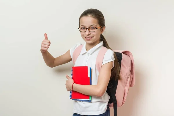 女孩学校学生, 用背包和笔记本显示竖起大拇指标志, 背景明亮的墙壁在学校 — 图库照片