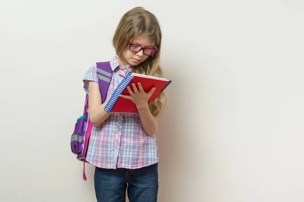 Niña estudiante de primaria con gafas con una mochila escribiendo en su cuaderno. Fondo brillante pared de la escuela — Foto de Stock