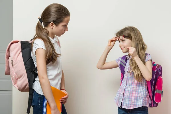 การสื่อสารของเด็กผู้หญิงสองคนที่โรงเรียน เด็กนักเรียนที่มีกระเป๋าเป้สะพายหลัง ผนังด้านหลังสดใสที่โรงเรียน — ภาพถ่ายสต็อก