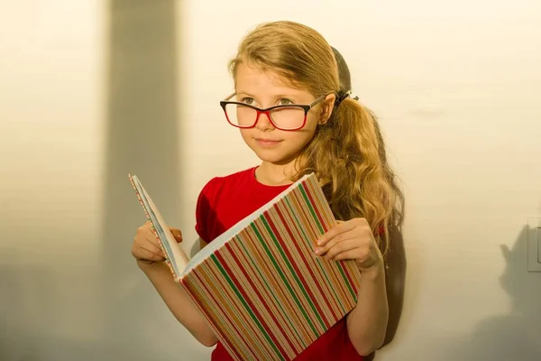 Niña niña estudiante de primaria con gafas está sosteniendo un libro de texto y sonriendo soñadoramente mirando hacia adelante —  Fotos de Stock