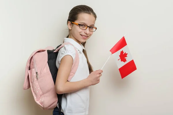 Studentessa 10 anni in t-shirt bianca con uno zaino che tiene la bandiera del Canada, sfondo parete luminosa nella scuola — Foto Stock