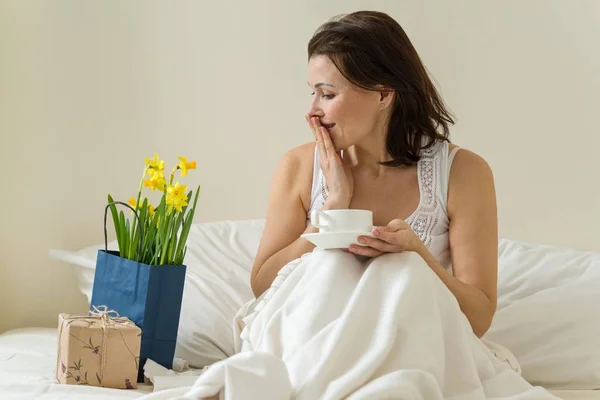 Attraktiva lycklig mellersta år kvinna kvinnliga är nöjd med gåva, bukett blommor som sitter på morgonen i sängen med en kopp kaffe. — Stockfoto