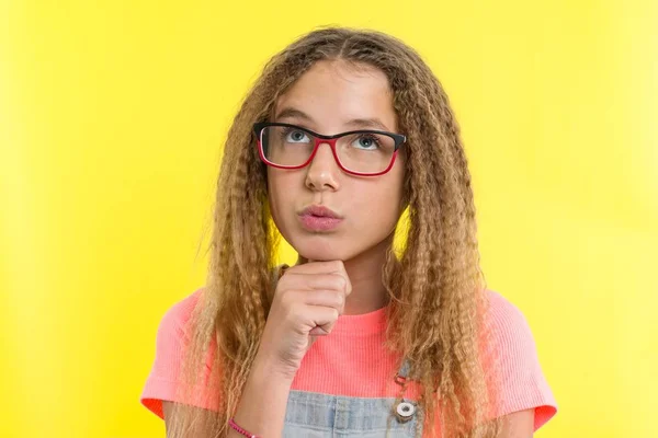 Söt flicka 12-13 år gammal blondin med lockigt hår med glasögon, ser tonföljd undan, tänker på skolan. Ansiktsuttryck koncept, gul bakgrund — Stockfoto